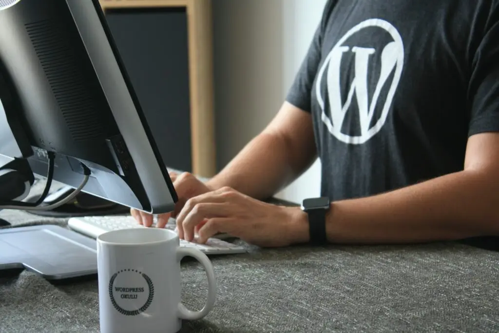 Person arbeitet am Computer mit WordPress-T-Shirt; Tasse mit „WordPress Okulu“ im Vordergrund